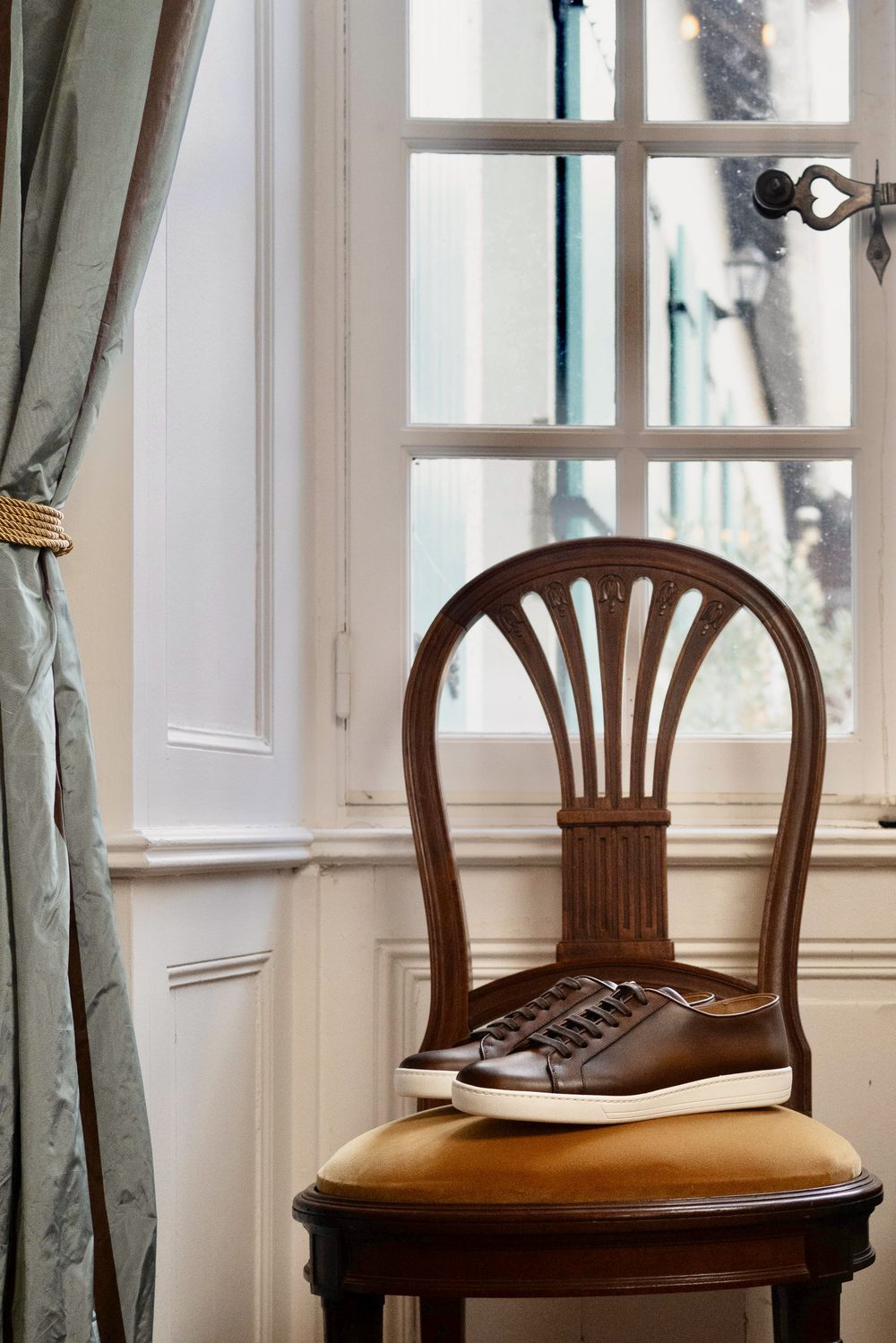 A pair of brown leather sartorial sneakers sits on a wooden chair with a yellow cushion.