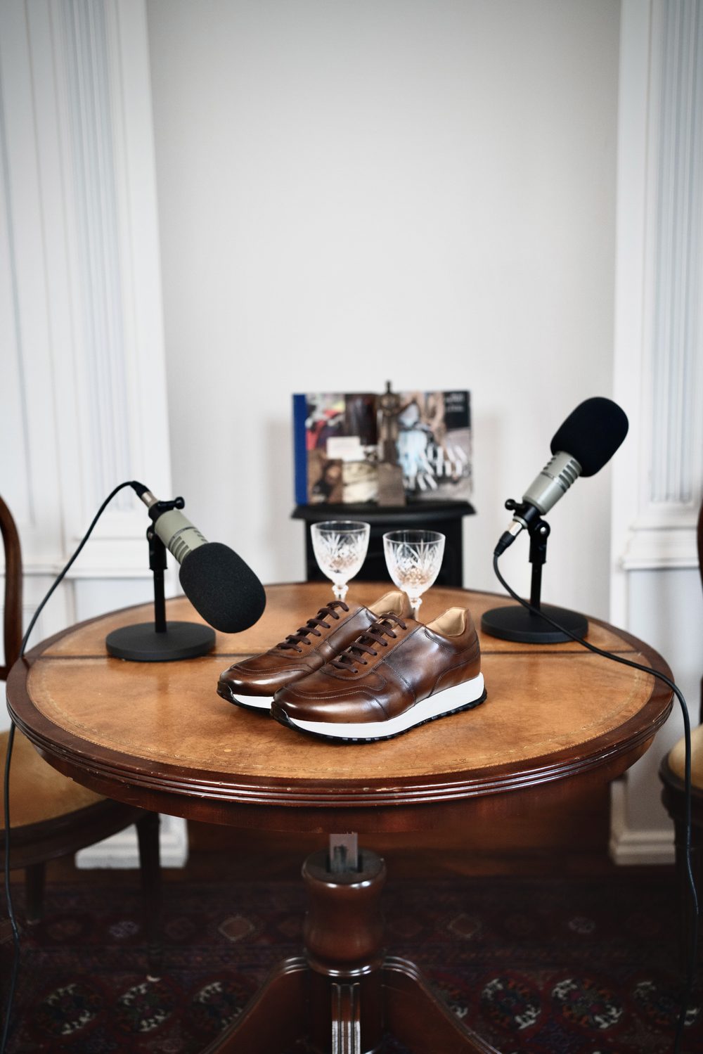 A pair of brown leather sartorial sneakers with white soles sits on a wooden table between two microphones.