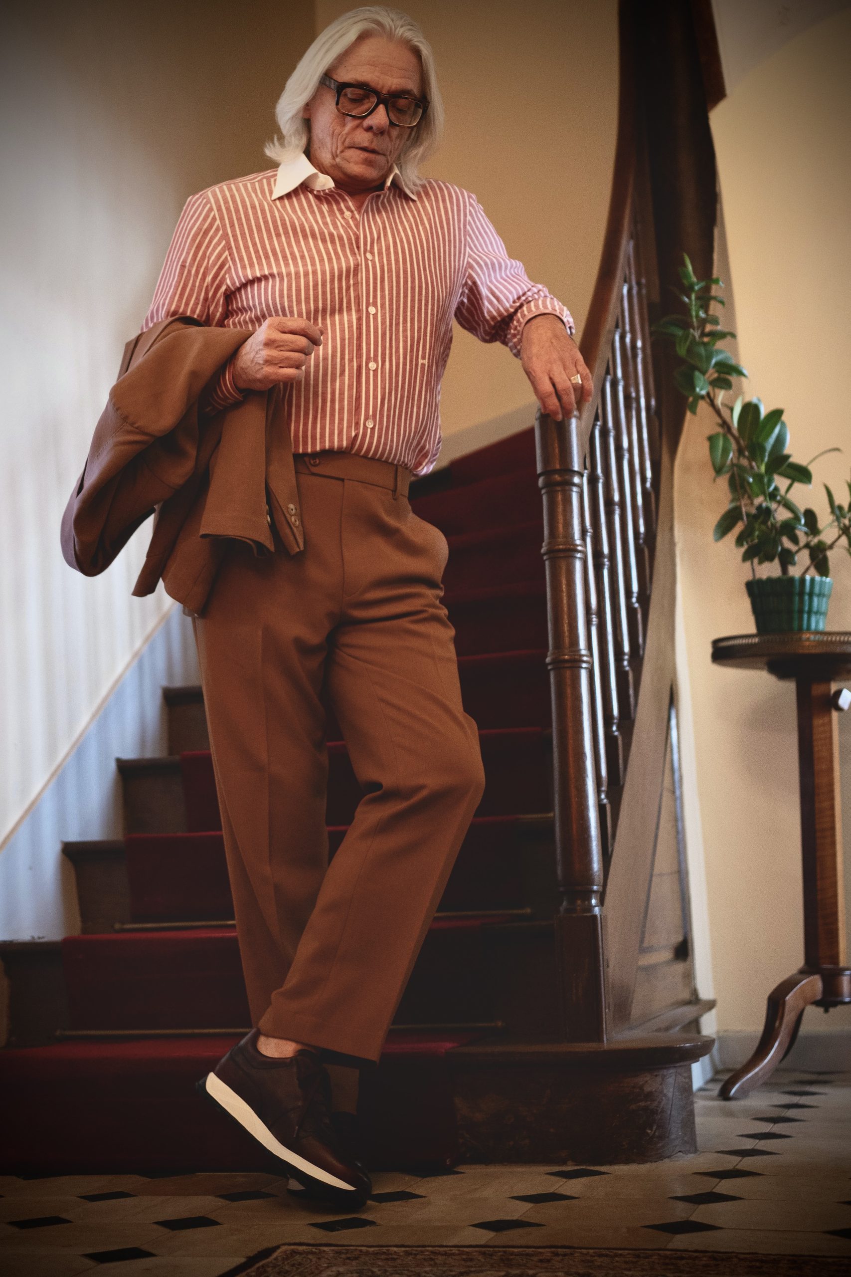 A man with long gray hair wears a striped red and white shirt, brown trousers, and dark sneakers while walking down a staircase.