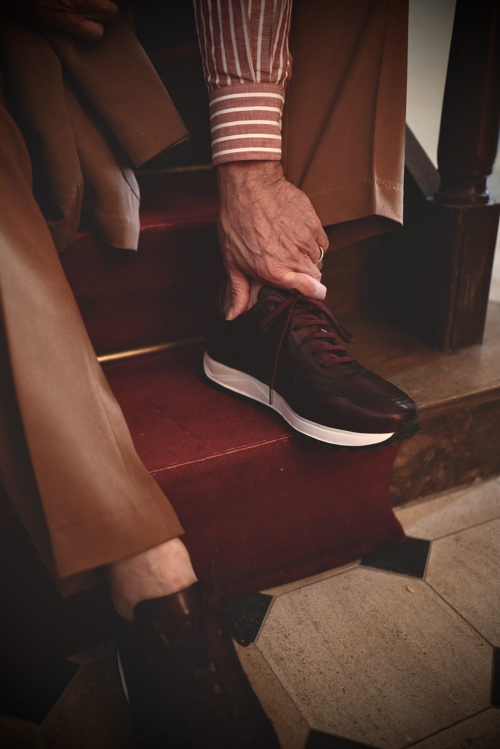 A close-up of a hand tying the laces on a dark brown sartorial sneaker with a white sole.