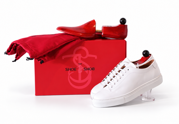 A red shoe box with a logo, a white sneaker, and a red shoe bag, illustrating how to properly store dress shoes.