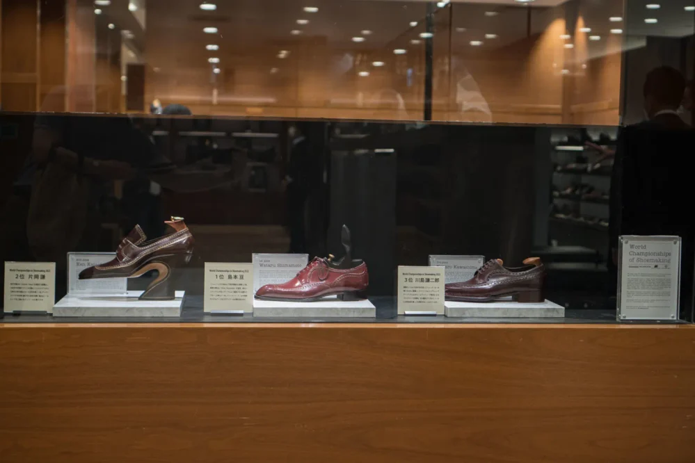 Three pairs of brown shoes displayed in a glass case, with labels indicating their rankings in the World Championships of Shoemaking 2027.