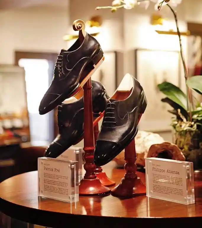Two pairs of elegant black dress shoes are displayed on wooden stands with name plaques for Patrick Frei and Philippe Atienza.
