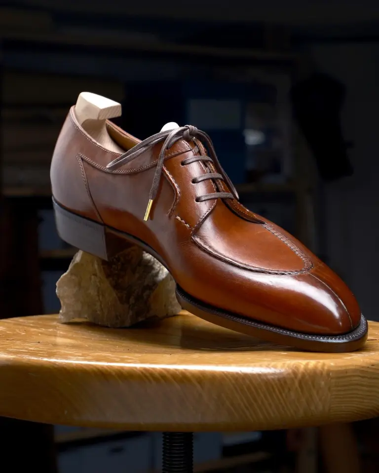 A polished brown leather shoe with a unique design and laces, displayed on a wooden surface.