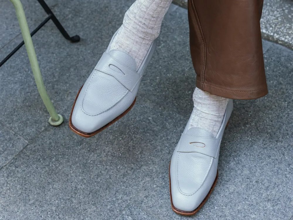 A pair of light gray leather loafers worn with cream-colored socks and brown pants.