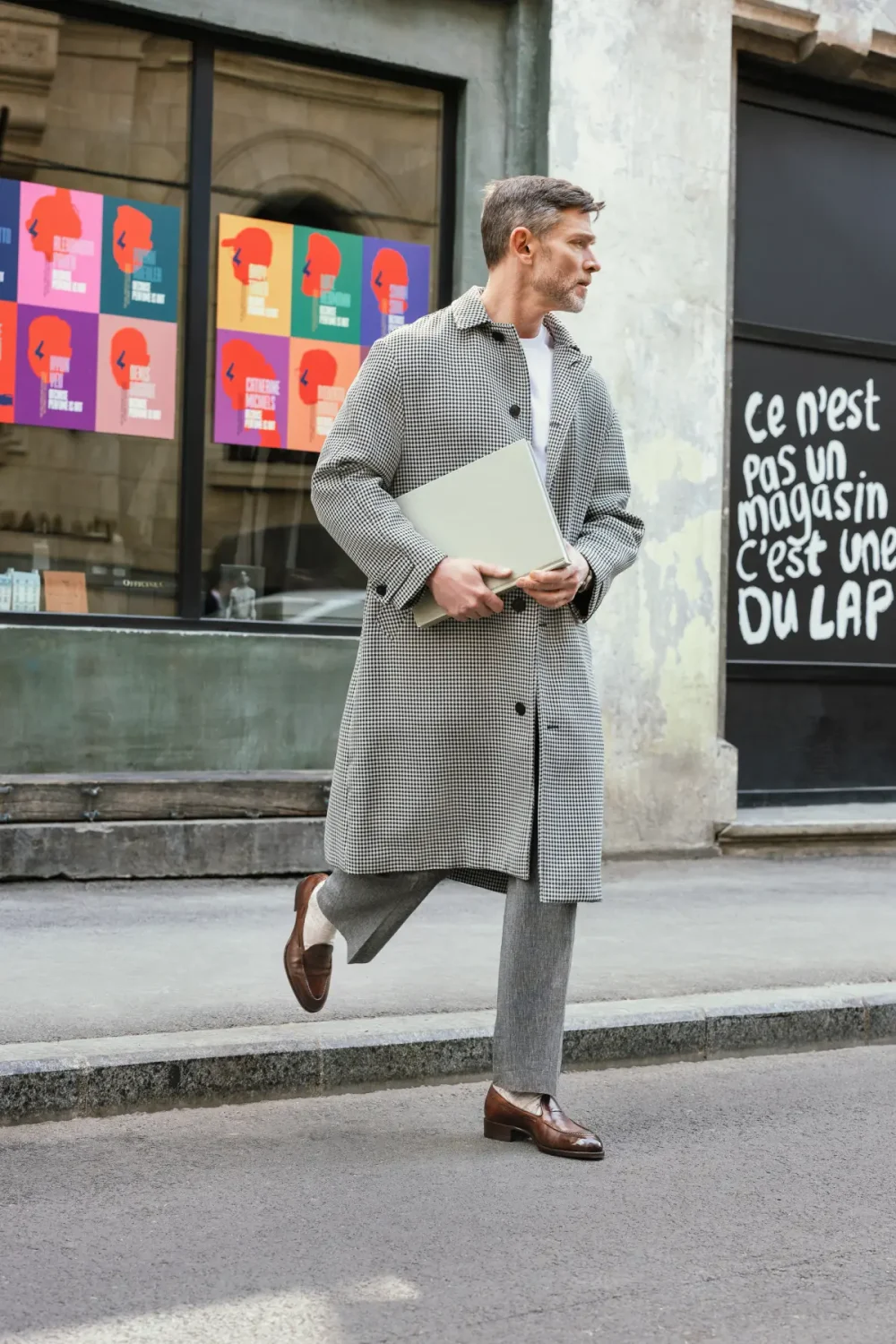 A man in a gray houndstooth coat and gray pants walks while holding a light-colored laptop.
