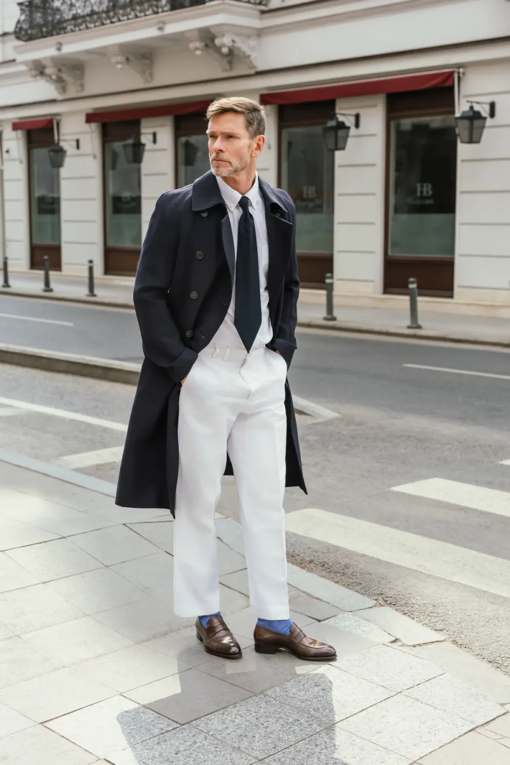 A man in a navy coat, white trousers, and brown shoes stands confidently with his hands in his pockets.