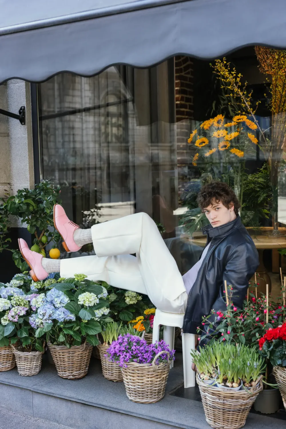 A young man in a black leather jacket and white pants sits with his legs crossed, showcasing pink shoes against a backdrop of colorful flowers in woven baskets.