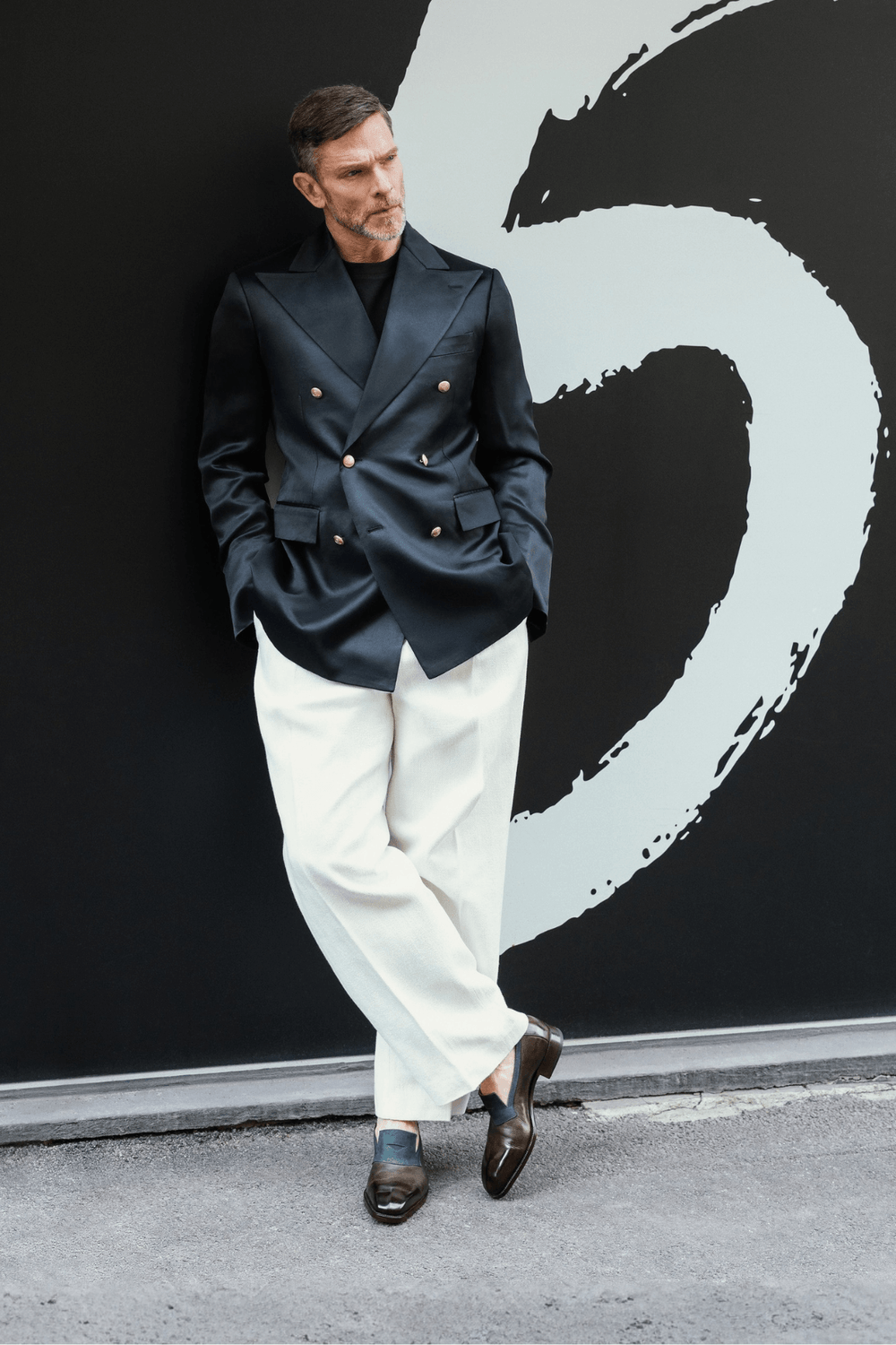 A man in a dark blue double-breasted blazer and white trousers stands with his hands in his pockets, wearing brown shoes with a blue sock.