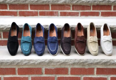 A row of eight pairs of suede loafers in black, teal, navy, brown, and cream, featuring a bow detail on each pair.