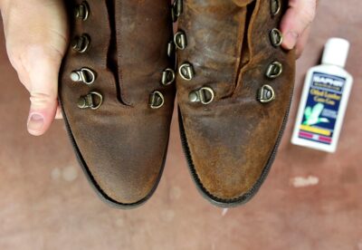 A pair of worn brown leather boots with metal eyelets, held in hands, alongside a bottle of Saphir Oiled Leather Cream.